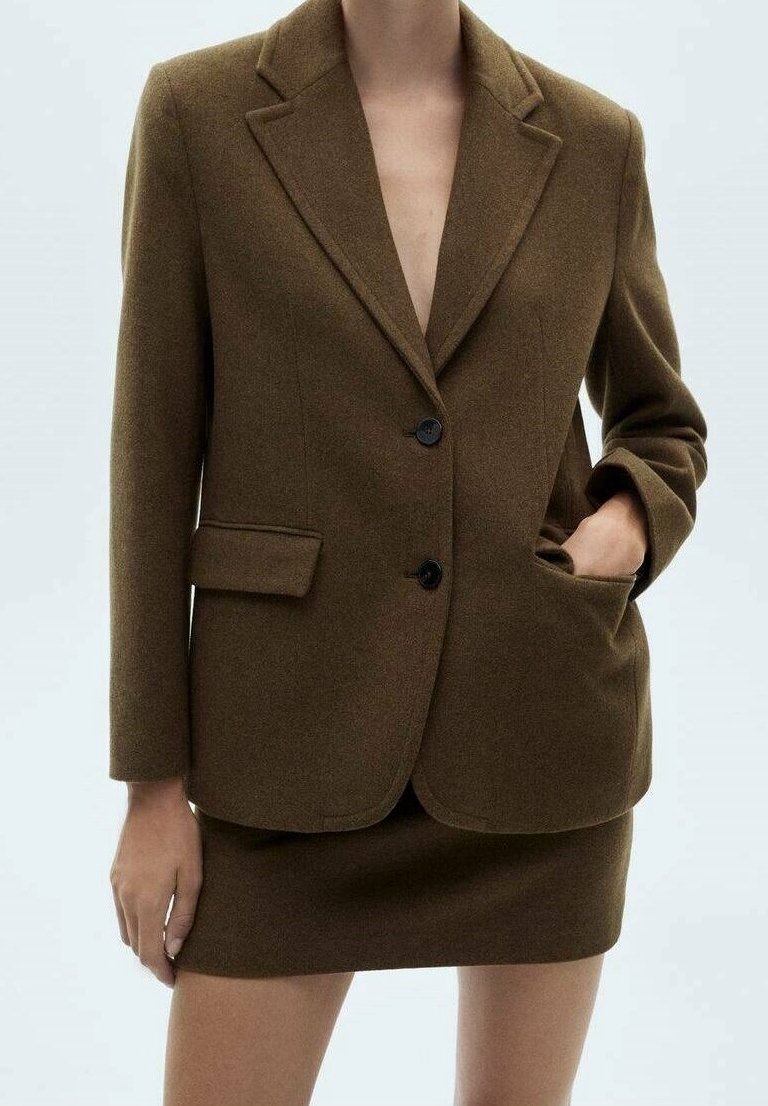 Femme portant un blazer marron à deux boutons avec poches avant, assorti à une mini-jupe marron sur un fond uni.
