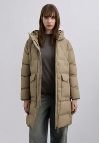 Abrigo beige acolchado con capucha, con diseño acolchado, bolsillos delanteros y botones de presión. Lleva puesto un suéter gris oscuro y vaqueros.