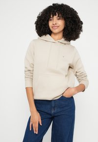 Μπεζ φούτερ με κουκούλα που σφίγγει με κορδόνι, μπροστινή τσέπη και ριμπ μανσέτες. Συνδυάζεται με μπλε τζιν παντελόνι με φαρδιά μπατζάκια. Απλός σχεδιασμός με ελάχιστες λεπτομέρειες.