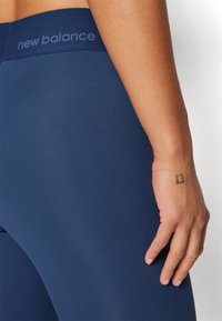 Primer plano de la mano derecha con un tatuaje de "13" descansando cerca de la cadera, usando leggings de New Balance azul marino con el logo en la cintura.