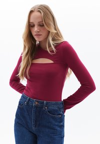 Pitkähihaisessa syvän burgundinvärisessä paidassa on etupuolella leikkaus, ja se on yhdistetty korkeavyötäröisiin sinisiin farkkuihin, joissa on napit yksityiskohtina.