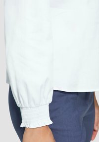 Weiße Bluse mit gerafften Manschetten, die über einen elastischen Saum verfügt. Getragen mit navyblauen, enganliegenden Hosen. Nahaufnahme hebt das Design der Ärmel hervor.