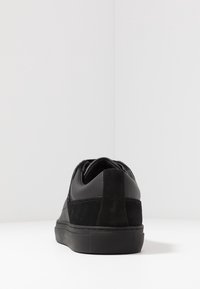 Musta tennari, joka on valmistettu nahasta ja mokkanahasta, jossa on teksturoitu takapuoli ja sileät sivut, litteällä kumipohjalla ja minimalistisella muotoilulla.