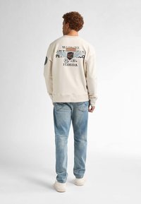 Felpa crema con stampa grafica sulla schiena, abbinata a jeans in denim azzurro chiaro. L'outfit presenta polsini a coste e una vestibilità rilassata.