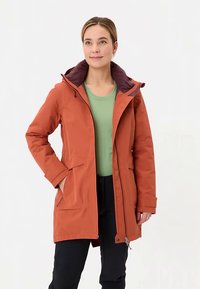 Waterdichte oranje jas met een bordeauxrode capuchon, twee voorzakken en een ritssluiting, gecombineerd met een groen shirt en zwarte broek.