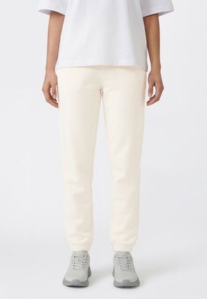 Osoba ubrana w jasnobeżowe spodnie dresowe, szare trampki i białą koszulkę z krótkim rękawem, stojąca na tle jednolitego jasnego tła.