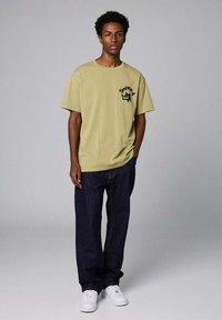 T-shirt verde oliva chiaro con logo grafico nero sul lato sinistro del petto, abbinato a jeans a gamba larga blu scuro e sneaker bianche.