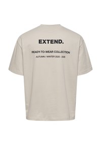 T-shirt beige con scollatura rotonda, maniche corte e testo sul retro: "EXTEND. COLLEZIONE PRONTA DA INDOSSARE AUTUNNO / INVERNO 2025-2026" in nero.