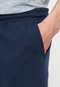 Main partiellement dans la poche d'un pantalon de survêtement bleu marine porté avec une chemise rayée rentrée.