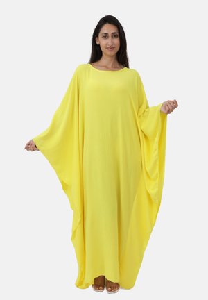 Mujer con un vestido suelto, amarillo brillante y hasta el tobillo, con mangas anchas, de pie con los brazos extendidos para mostrar la prenda.