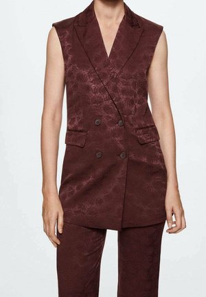 Mouwloos dubbelgeknoopt bordeauxblazer met subtiel bloemmotief, gedragen met bijpassende broek op een persoon tegen een effen achtergrond.