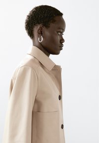 Profil af en person med kort krøllet hår iført sølvfarvede hoop øreringe og en beige knapjakke mod en ensfarvet lys baggrund.