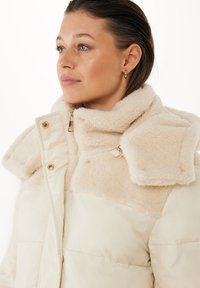 Beige pufferjacka med plyschshearlingdetaljer på kragen och axlarna. Har en framzip och detaljer i guld.