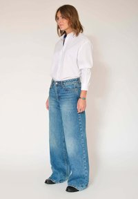 Jean bleu clair ample avec une texture délavée, associé à une chemise blanche boutonnée. Des chaussures noires complètent la tenue.