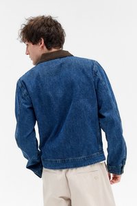 Ung mand med krøllet hår iført en blå denimjakke med en mørkebrun krave og beige bukser, som vender væk mod en hvid baggrund.