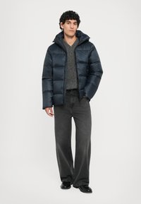 Marineblaues Puffermantel mit gestepptem Design, Kapuze und frontalem Reißverschluss. Getragen über einem grauen Pullover und kombiniert mit lässigen schwarzen Jeans und schwarzen Schuhen.