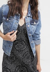 Jeansjacke mit taillierter Passform, hellblauer Farbe, Knopfverschlüssen, zwei Brusttaschen und aufgerollten Ärmeln über einem schwarz gemusterten Kleid.