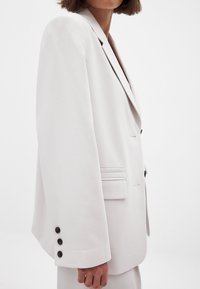 Gai pelēks žakets ar pielāgotu piegriezumu, vienpogu dizainu, atlocītām atloks un trim tumšajiem pogām pie piedurknēm. Smalka tekstūra un minimālas detaļas.