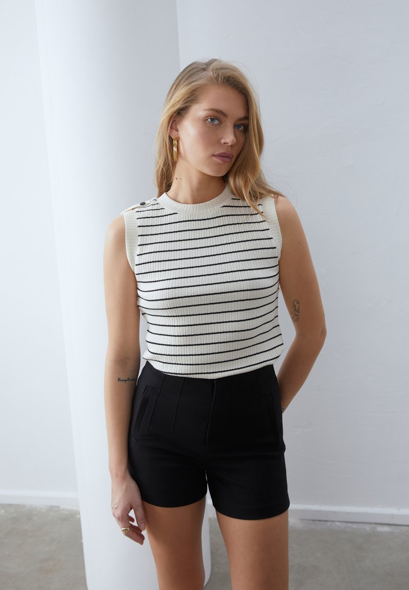 Gestreifter, gerippter ärmelloser Oberteil in Creme und Schwarz, mit Schulterknöpfen; kombiniert mit hoch taillierten schwarzen Shorts mit vorderen Falten.