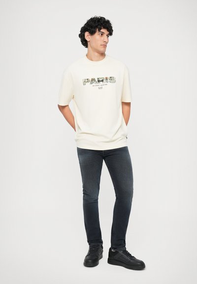 Vitt bomullströja med grafiken "PARIS" i grönt och brunt, kombinerad med mörka jeans och svarta sneakers, står med händerna i fickorna.