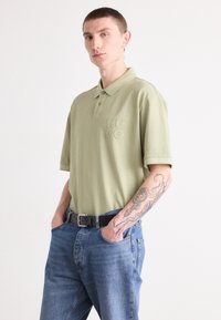 Polo shirt vert olive à manches courtes, logo "HUGO" brodé sur la poitrine, associé à un jean en denim bleu et une ceinture noire.