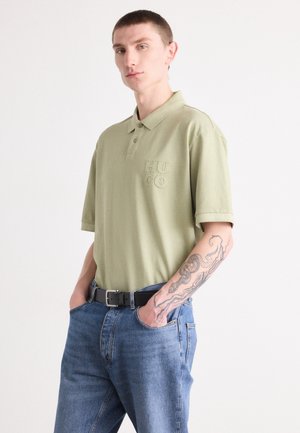Olive green polo shirt with short sleeves, embroidered "HUGO" logo on the chest, paired with blue denim jeans and a black belt.