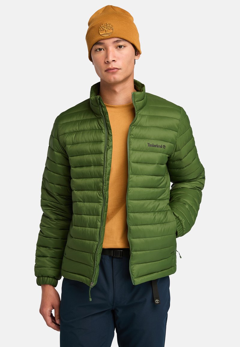 Groene pufferjack met horizontale stiksels, ritssluiting aan de voorkant en opstaande kraag. Gecombineerd met een mosterdkleurig shirt en een marineblauwe broek, gepresenteerd met een beanie.