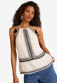 Top alb cu bretele, brodat în negru și cu accente de ciucuri, având un corset ajustat și o fustă evazată, purtat cu pantaloni scurți din denim.
