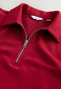 Nærbillede af en rød zip-up fleecekrave skjorte med sølv lynlås og et mærket hvidt tag indeni kraven.