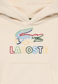 Sudadera con capucha con logotipo de cocodrilo bordado multicolor y texto "LACOSTE" en tela beige sobre el bolsillo tipo canguro.