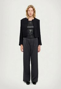 Mustat rypytetty takki, joka on pukeutunut kiiltävän mustan topin päälle, yhdistettyinä löysään harmaaseen housuun ja mustiin kenkiin.