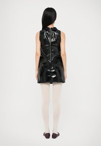 Γυναίκα με μακριά μαύρα μαλλιά που φοράει ένα αμάνικο μαύρο μίνι φόρεμα από λακαρισμένο δέρμα, λευκές καλσόν και μαύρες επίπεδες παπούτσια, στέκεται με την πλάτη στραμμένη.