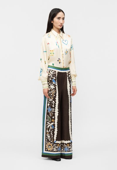 Γυναίκα με εκρού  μπλούζα με πολύχρωμα σχέδια και μαύρο παντελόνι με φαρδιά πόδια, διακοσμημένο με περίπλοκα μπλε, πράσινα και καφέ λουλουδένια μοτίβα.