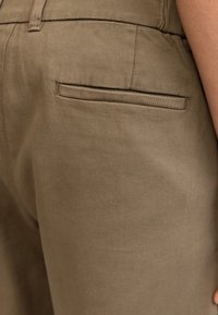Brown fabric pants featuring a smooth texture, slim fit, and a single back pocket with a subtle stitching detail.
