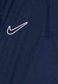 Mørkeblå treningstopp med glidelås i halsen og en hvit brodert Nike-logo på brystet. Materialet har et glatt, teksturert utseende.