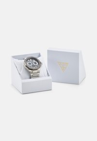 Reloj de tono plateado con una esfera gris texturizada, funciones de cronógrafo y una correa de malla metálica. Presentado en una caja blanca con un logo dorado.