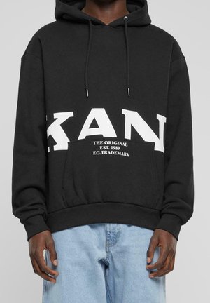 Zwarte capuchontrui met grote witte tekst "KAN" en kleinere witte tekst "THE ORIGINAL EST. 1989 EG.TRADEMARK," gedragen met lichtblauwe spijkerbroek.