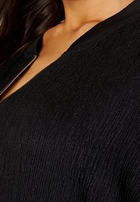 Cardigan nero texture con un pattern a coste, dotato di zip frontale e colletto liscio.
