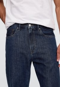 Ciemnoniebieskie dżinsy z denimu o prostym kroju, z pięcioma kieszeniami i widocznymi detalami przeszyć, zapinane na metalowy guzik w pasie.