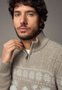 Hellbrauner gestrickter Pullover mit einem Reißverschlusskragen, der weiße Schneeflockenmuster und gerippte Bündchen aufweist. Weiche Textur und detailliertes Design.
