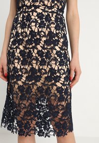 Femme portant une robe midi beige avec une superposition en dentelle à fleurs bleu marine et du vernis à ongles rouge, debout devant un fond uni.