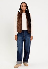 Femme avec de longues tresses portant un gilet en fausse fourrure marron sur un pull crème, un jean large à revers et des baskets beiges, se tenant devant un fond blanc.