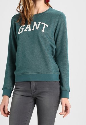 Femme portant un sweat-shirt GANT bleu sarcelle et un jean gris foncé, debout, les bras détendus le long du corps, devant un fond uni.