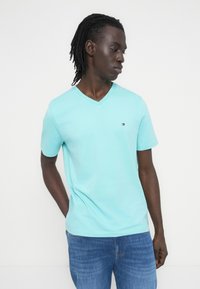 Τυρκουάζ μπλουζάκι με V λαιμόκοψη από απαλό βαμβάκι, με κοντά μανίκια. Μικρό κεντημένο λογότυπο στο στήθος. Συνδυάζεται με μπλε τζιν.