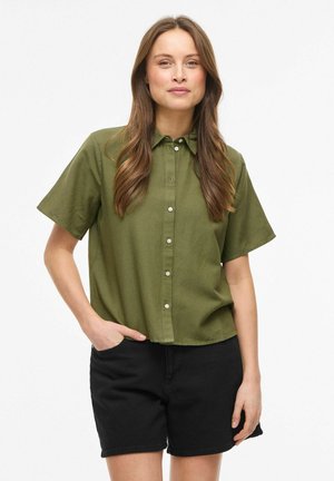 Kvinne med langt brunt hår iført en olivengrønn kortermet skjorte med knapper og svarte shorts, stående med en hånd i lommen.