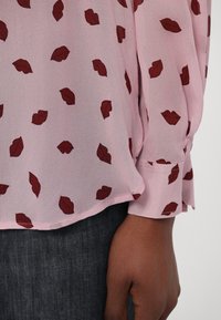 Pink blouse featuring a lightweight, sheer fabric with a repeated pattern of dark red lips. Includes long sleeves with a cuff detail.