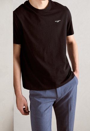 Camiseta negra de algodón con mangas cortas y cuello redondo, con un logo blanco en la parte superior izquierda del pecho. Combinada con pantalones azul claro.