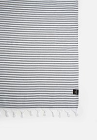Textil a rayas en gris y blanco con un acabado texturizado. Presenta borlas decorativas blancas a lo largo del borde y una etiqueta negra.