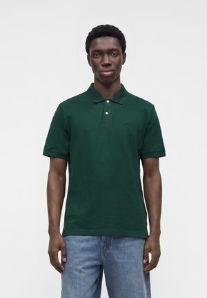 Mand står fremadvendt iført en mørkegrøn poloshirt og blå jeans mod en ensfarvet lys baggrund.