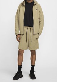 Beige zip-up huvtröja med svarta detaljer, tillsammans med beige shorts med dragsko och sidofickor. Svart T-shirt, strumpor och träningsskor.